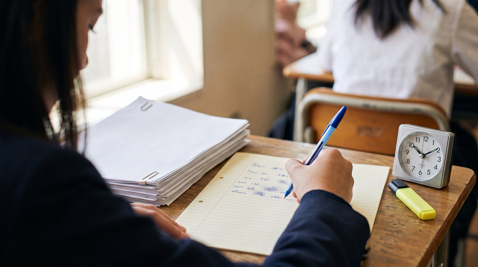 Close-up of a senior student's hand writing kanji on lined exam paper beside a clock and past papers